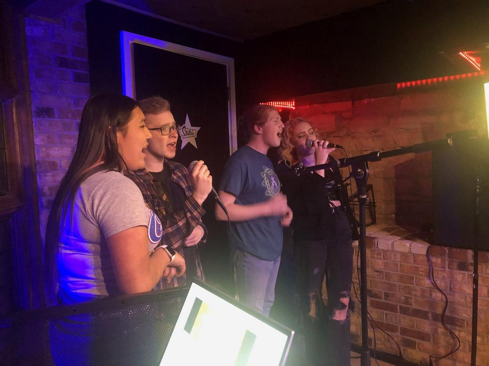 four singers sharing microphones in front of a lyrics screen