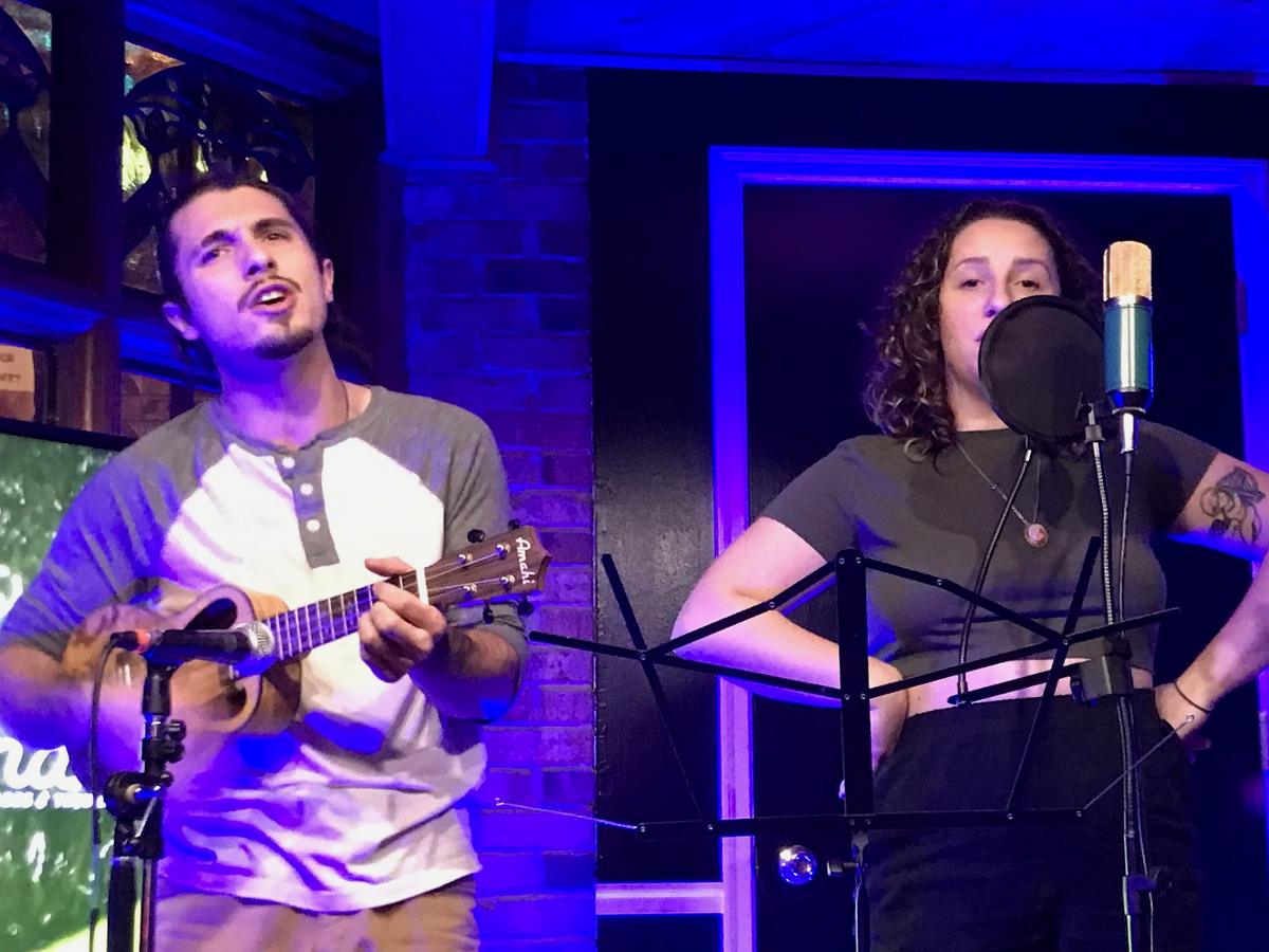a singer at a studio microphone on a blue-lit stage, accompanied by a ukulele player
